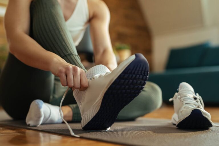 Person sitting on a mat and tying the laces of a white athletic shoe, preparing for a workout.